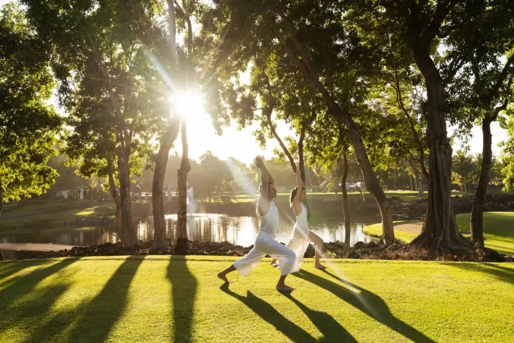 Zwei weiß gekleidete Personen praktizieren Yoga in der Krieger-1-Pose vor dem idyllisch grünen Hintergrund des Constance Belle Mare Plage auf Mauritius