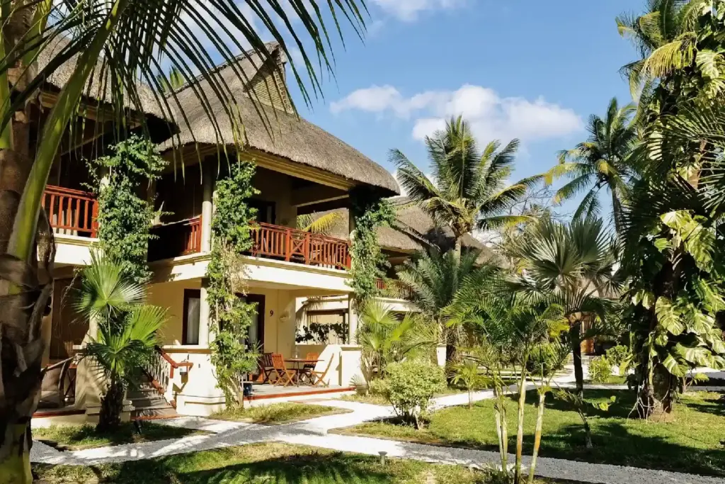 Ein mehstöckiger, palmgedeckter Bungalow mit Terrasse und Balkon, davor Palmen und viel Grün - buchen Sie Ihre Unterkunft auf der Trauminsel Mauritius mit Fenners Reisen