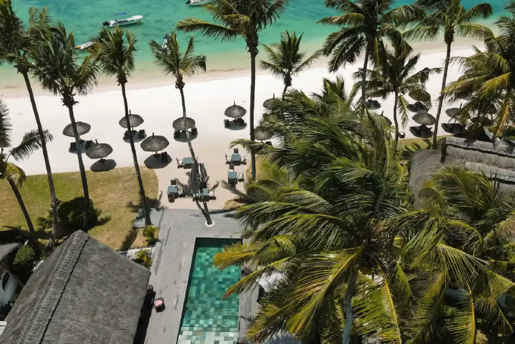 Sicht von oben auf einen türkisen Pool, Sonnenschirme am weißen Palmenstrand und das tiefblaue Meer - erleben Sie mit Fenners Reisen die landschaftlichen Kontraste der Insel Mauritius!