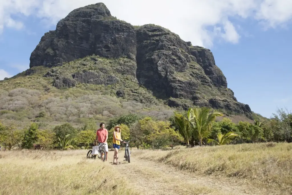 Ein Paar schiebt seine Räder durch grüne Wiesen vor dem Hintergrund des ikonischen Le Morne Brabant Berges. Mauritius hat noch viel mehr zu bieten als Sonne und Strand! Katharina Fenners berät Sie individuell und persönlich.
