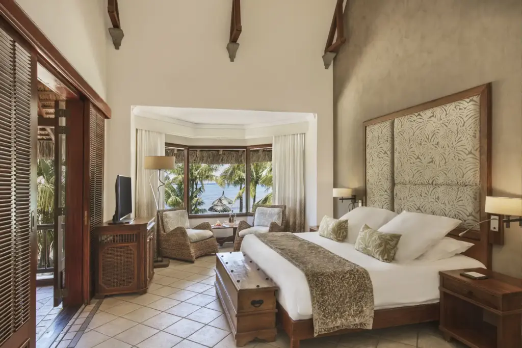 Eine luxuriöse Hotelsuite mit großen Fenstern, aus denen der Blick auf einen palmgesäumten Strand geht - so komfortabel übernachten Gäste des Dinarobin Beachcomber Golf Resort & Spa, buchbar bei Fenners Reisen