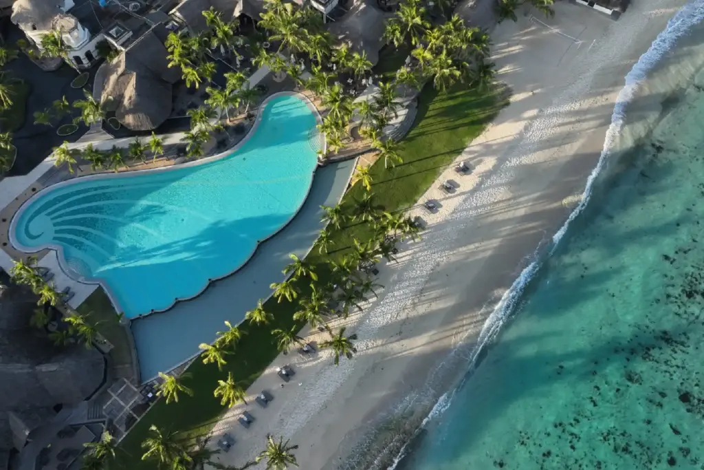 Blick von oben auf einen wellenförmigen Pool an einem menschenleeren Sandstrand, davor türkises Meer - ein Aufenthalt im Dinarobin Beachcomber Golf Resort & Spa vereint das Beste, was die Trauminsel Mauritius zu bieten hat und kann bei Fenners Reisen gebucht werden