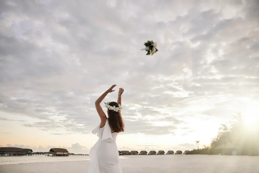 Eine Braut steht im Sonnenuntergang auf dem leeren Strand einer maledivischen Insel und wirft den Brautstrauß. Am Horizont reihen sich Wasservillen wie Perlen entlang. Buchen Sie Ihre Hochzeit und Flitterwochen auf den Malediven bei Fenners Reisen!