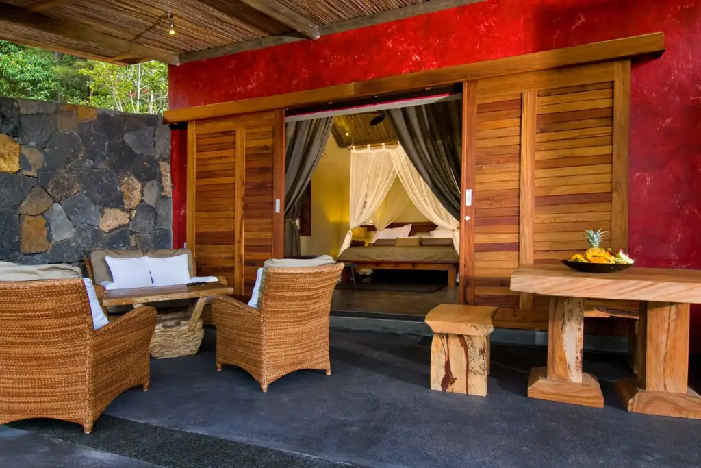 Eine rustikal möblierte Terrasse vor einem rot gestrichenen Gebäude, dessen bodentiefe Fensterfront von charmanten Holzfensterläden begrenzt wird. Buchen Sie einen naturnahen Aufenthalt auf Mauritius mitten im Nationalpark bei Fenners Reisen!