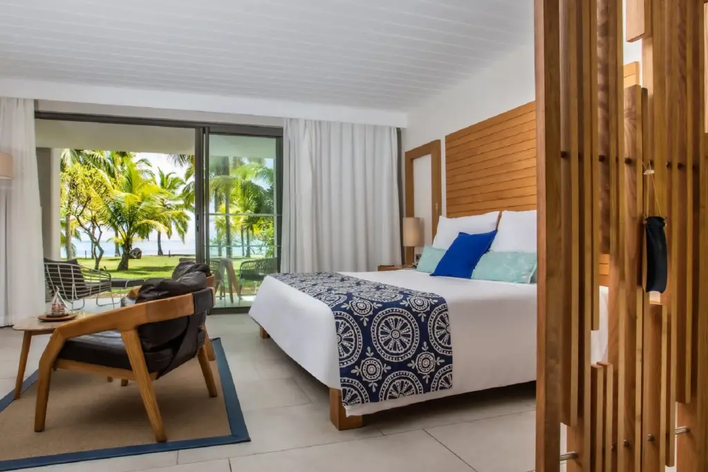 Ein schick gestaltetes Hotelzimmer mit viel Holz und bodentiefen Fensterfronten, aus denen der Blick ins Grüne und bis zum Ozean schweift - buchen Sie bei Fenners Reisen einen maßgeschneiderten Traumurlaub auf Mauritius!