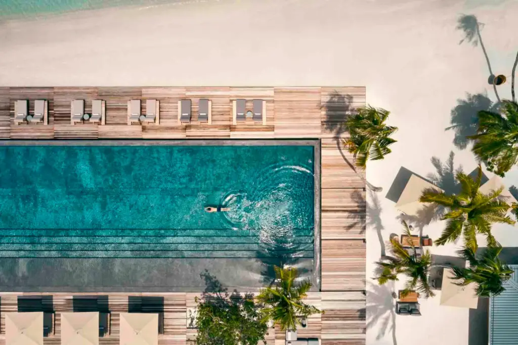 Blick auf einen Pool aus der Luft, umgeben von stilvollen Outdoor-Möbeln und strahlend weißem Sand. Katharina Fenners ist Spezialistin für Luxusreisen auf die Malediven und berät Sie individuell und persönlich!