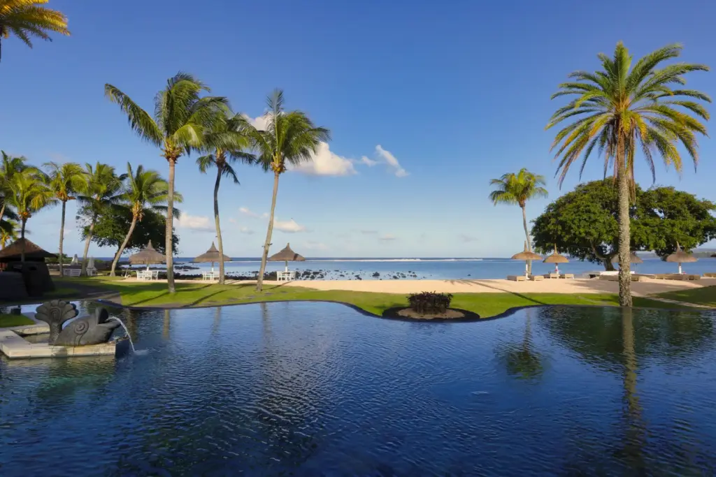 Blick über einen großen, tiefblauen Pool und den dahinterliegenden Sandstrand am Indischen Ozean. Buchen Sie eine exklusive Auszeit auf Mauritius! Katharina Fenners berät Sie individuell und persönlich.