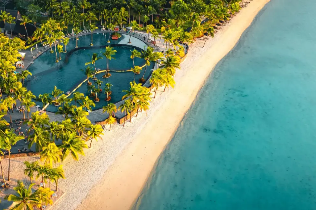Blick von oben auf den leuchtenden Strand und den tiefblauen Ozean direkt vor dem von Palmen umstandenen Hauptpool des Trou aux Biches Beachcomber Golf Resort & Spa, buchbar bei Fenners Reisen!