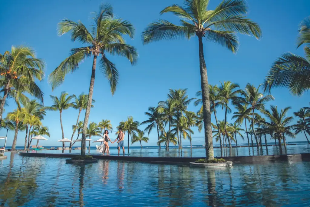 Ein Paar flaniert unter hohen Palmen zwischen dem Pool und dem Ozean des Trou aux Biches Beachcomber Hotels auf Mauritius. Katharina Fenners arrangiert Ihren Aufenthalt zu besten Konditionen!