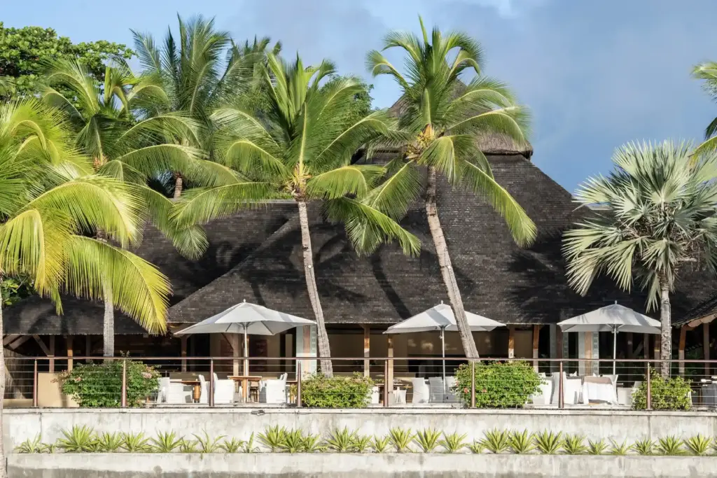 Hohe Palmen und einzelne Tische unter Sonnenschirme - so speist es sich direkt am Strand in einem der Restaurants des Trou aux Biches Beachcomber Golf Resorts & Spas, buchbar bei Fenners Reisen