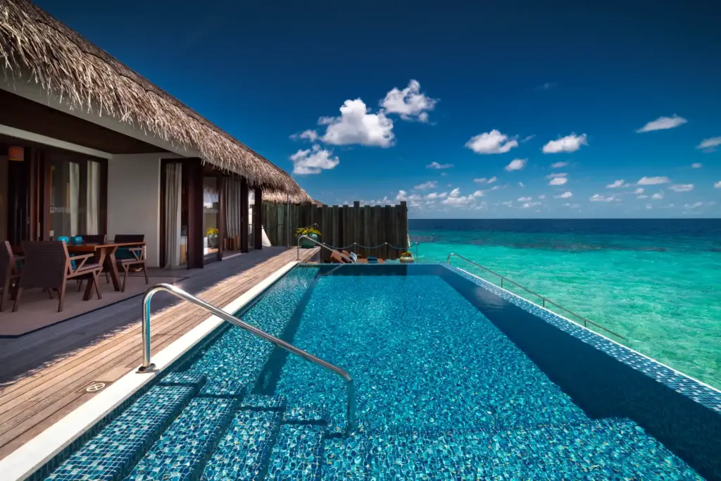 Glitzernder Infinity Pool vor dem türkisen Ozean, Sonnenliegen und ein Essbereich im Schatten - das bietet der großzügige Außenbereich einer Sunrise Water Pool Villa des Velaa Private Island Luxusresorts auf den Malediven, buchbar bei Fenners Reisen