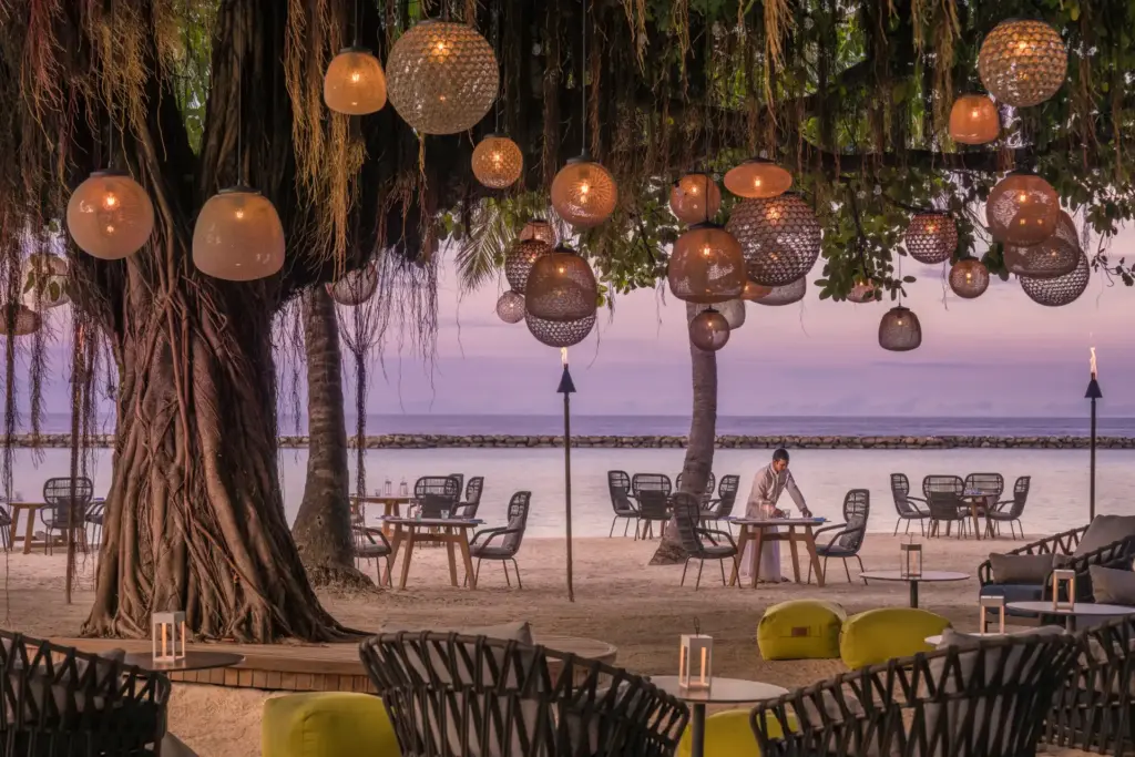 Festliche Lampen schmücken ein Strand-Restaurant in der rosa schimmernden Abenddämmerung vor dem Four Seasons Resort Maldives at Landaa Giraavaru. Buchen Sie einen traumhaften Luxusurlaub auf den Malediven bei der Malediven-Expertin Katharina Fenners!