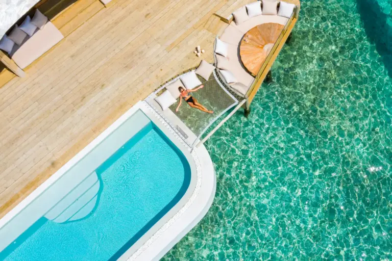 Blick aus der Vogelperspektive auf eine Frau, die in einem Hängenetz über dem Indischen Ozean entspannt, direkt neben dem eigenen Privatpool. Sie planen einen Luxusurlaub auf den Malediven? Katharina Fenners berät Sie individuell und organisiert den perfekten Aufenthalt für Sie.