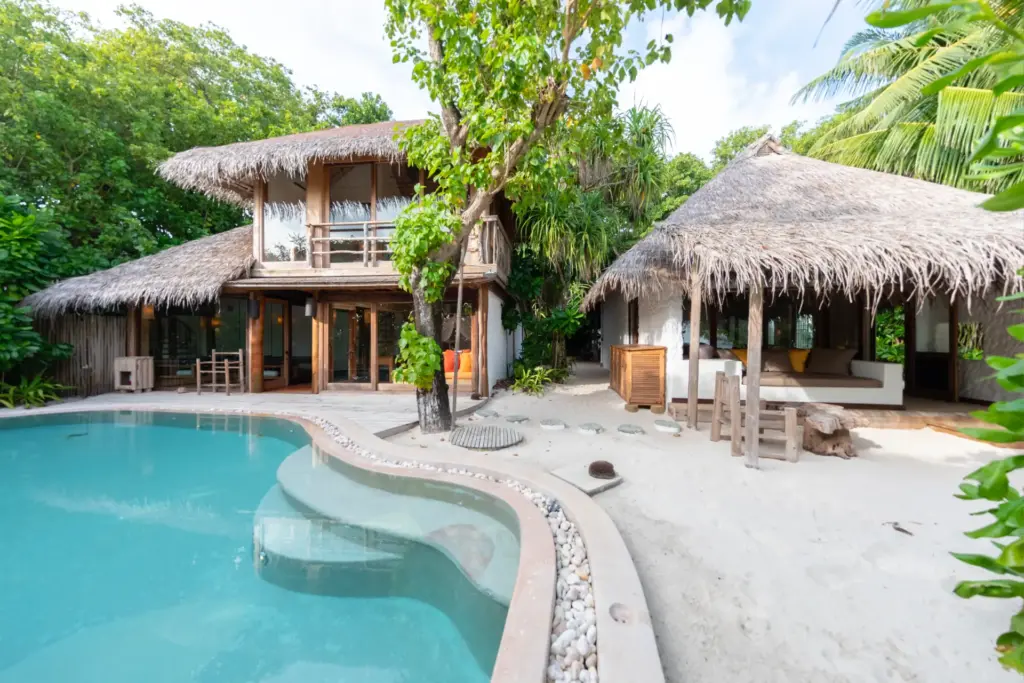 Ein wellenförmiger Pool vor zwei strohgedeckten Villen in üppiger tropischer Vegetation - ein Luxusurlaub auf den Malediven bringt Sie der Natur ganz nah, ohne Abstriche beim Komfort - Katharina Fenners kennt die besten Villen auf den Inseln am Indischen Ozean und berät Sie individuell!