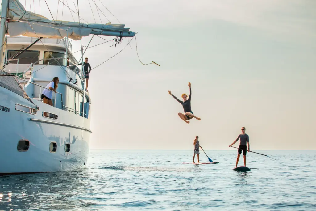 SUP-Fahrer umrunden eine Segelyacht im Indischen Ozean, während eine Urlauberin von Deck ins Wasser springt - unvergessliche Momente einer Luxusreise auf die Malediven, wie sie bei Fenners Reisen gebucht werden kann