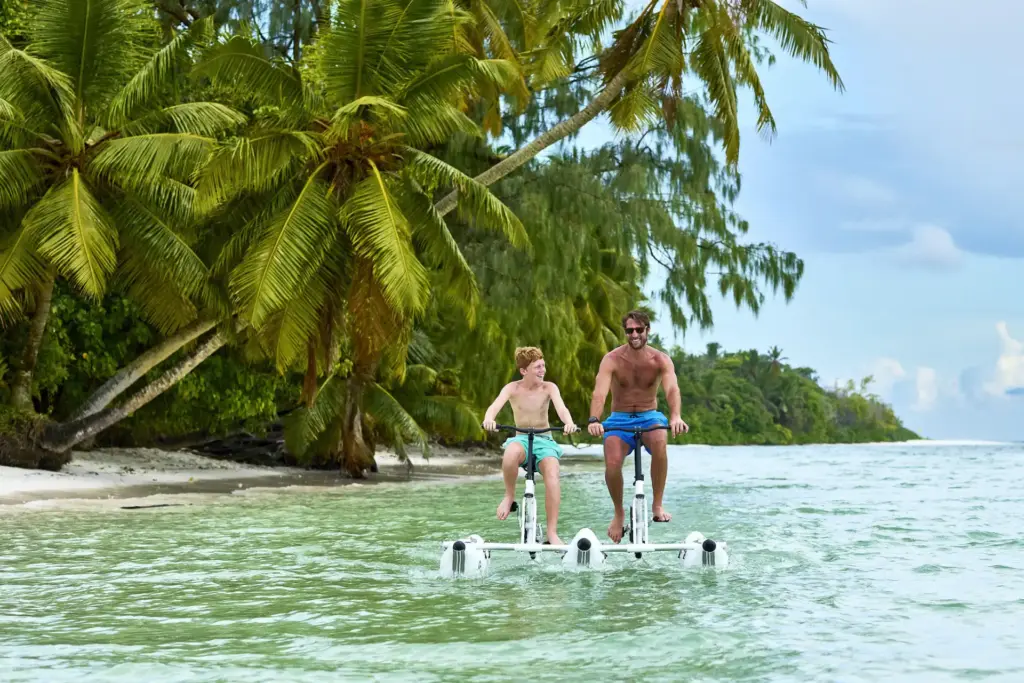 Vater und Sohn testen ein Wasserfahrrad im seichten Wasser des Indischen Ozeans, im Hintergrund beugen sich schwere Palmen über den weißen Sandstrand - eine Kulisse wie im Paradies. Katharina Fenners kennt die besten Resorts und Hotels für Ihren Familienurlaub auf den Seychellen und berät Sie individuell!