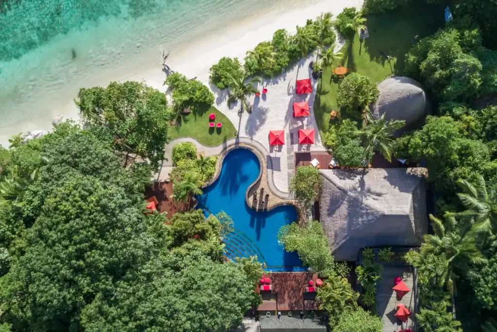 Vogelperspektive auf das Anantara Maia Seychelles Villas - rote Sonnenschirme umringen den wellenförmigen Pool, nur wenige Schritte vom weichen Sandstrand und dem türkisen Ozean entfernt. Ein unberührtes Paradies für Anspruchsvolle, buchbar für Ihre Seychellen Luxusreise bei Fenners Reisen.