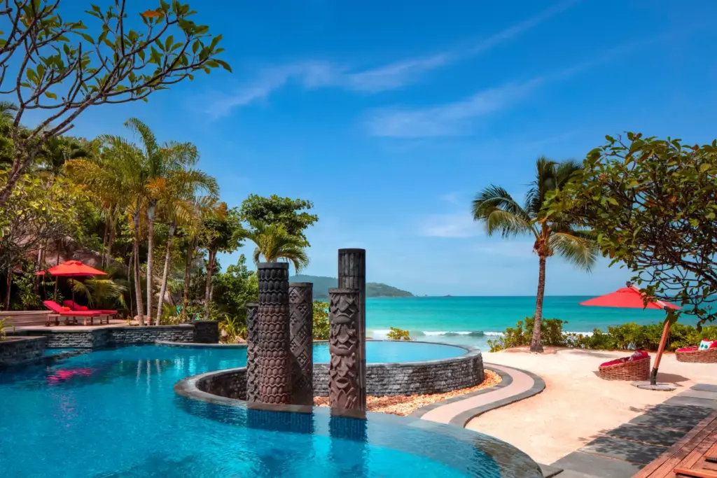 Um den geschwungenen Hauptpool des intimen Anantara Maia Seychelles Villas reihen sich imposante Skulpturen und rote Sonnenschirme und Liegen - buchen Sie einen Aufenthalt in einer der exklusiven Seychellen-Villen mit Butler-Service bei Fenners Reisen!
