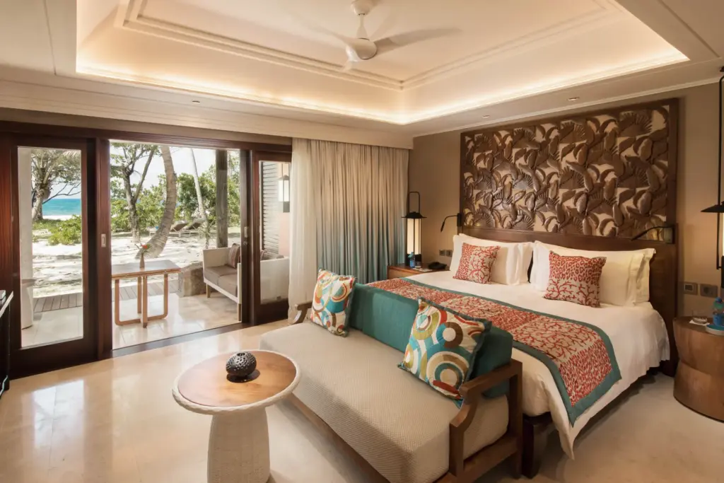 Bunte Akzente geben diesem Schlafzimmer einer Senior Suite einen modernen Look, während hinter den bodentiefen Fensterfronten die paradiesische Kulisse der Seychellen-Insel Praslin und der blau leuchtende Indische Ozean zu sehen sind. Ein Urlaub wie im Paradies, buchbar bei Fenners Reisen!