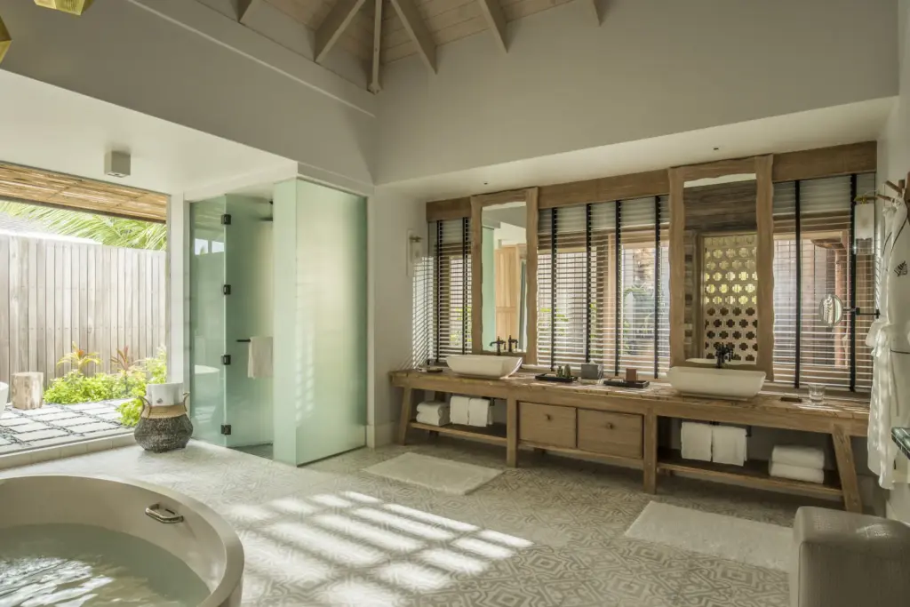 Ein weitläufiges Badezimmer mit doppeltem Waschtisch, kreisrunder Wanne und Outdoor-Dusche. Es gehört zu einer exklusiven Residenz auf einer intimen Privatinsel der Seychellen, buchbar für Ihren Seychellen Luxusurlaub bei Fenners Reisen!