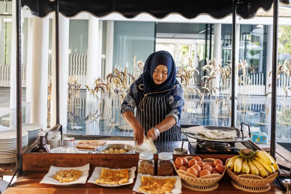 Eine Köchin in traditionell thailändischem Gewand serviert Köstlichkeiten am Frühstücksbuffet des Banyan Tree Krabi, buchbar bei Fenners Reisen