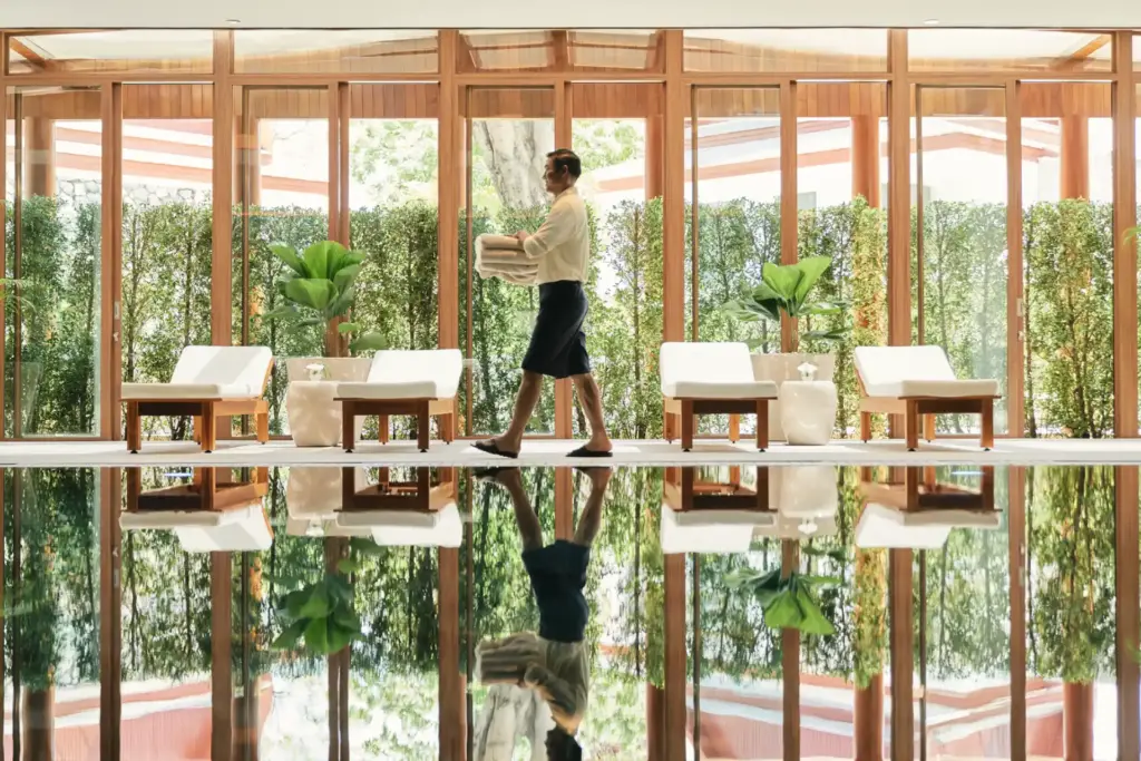Ein Angestellter des Chiva-Som Hua Hin spiegelt sich im Pool, während er vor der grünen Kulisse der umgebenden Natur Handtücher zu einer Liege bringt. So ikonisch wie dieses Foto ist auch der Aufenthalt im preisgekrönten Wellnessresort, buchbar bei Fenners Reisen.
