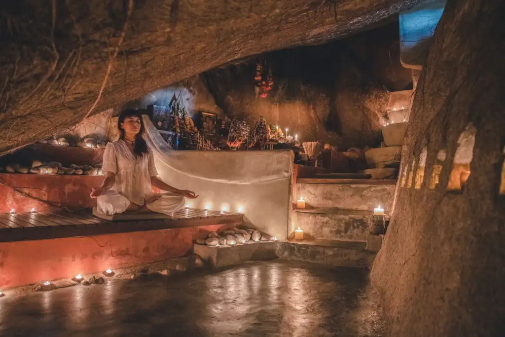 Eine Frau meditiert im flackernden Kerzenschein einer Höhle. Das preisgekrönte Wellness-Resort Kamalaya Koh Samui ist um diese jahrhundertealte Höhle herum gebaut und zehrt von deren besonderer Magie - kein Wunder, dass es ein echter Zufluchtsort für spirituelle Retreats ist. Katharina Fenners organisiert Ihren maßgeschneiderten Aufenthalt!