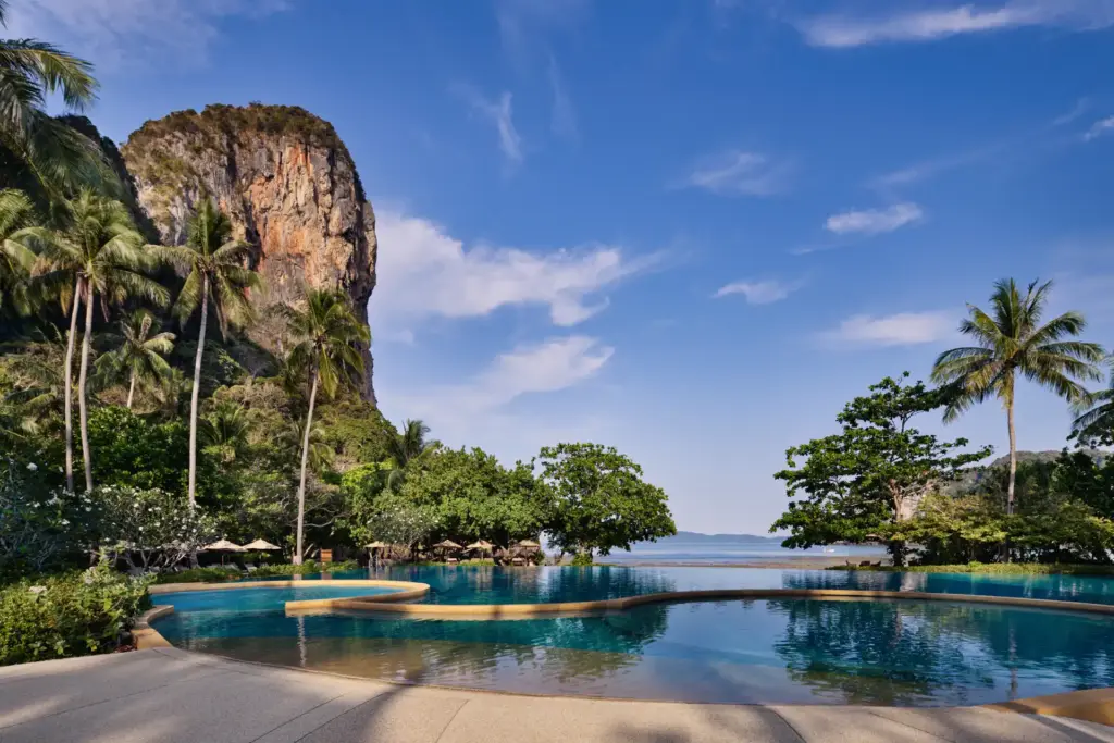 Der Hauptpool des Rayavadee Krabi erlaubt bezaubernde Blicke auf nahegelegene Felsprünge und den dahinter schimmernden Ozean. So atemberaubend ist die Kulisse Ihres Thailand Luxusurlaubs, maßgeschneidert organisiert von Fenners Reisen.