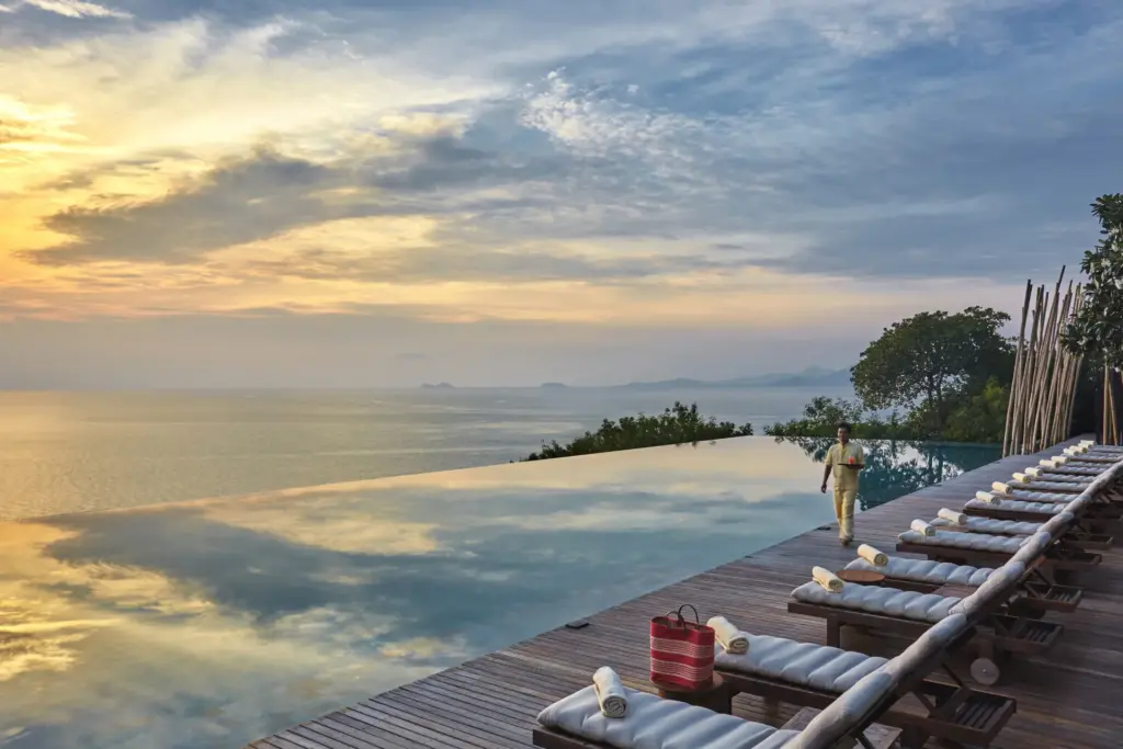Der Hauptpool nahezu olympischer Dimension leuchtet in den Farben des Sonnenuntergangs, während ein Angestellter Handtücher herbeibringt - eine der vielen erinnerungswürdigen Kulissen eines Thailand Luxusurlaubs im Six Senses Samui. Katharina Fenners organisiert den Aufenthalt!