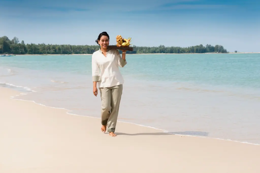 Eine Frau trägt lächelnd ein Tablett mit frischem Obst über den schier endlosen, feinsandigen Strand von Khao Lak. Sie sind auf der Suche nach den besten Resorts für einen exklusiven Strandurlaub in Thailand? Katharina Fenners berät Sie persönlich!