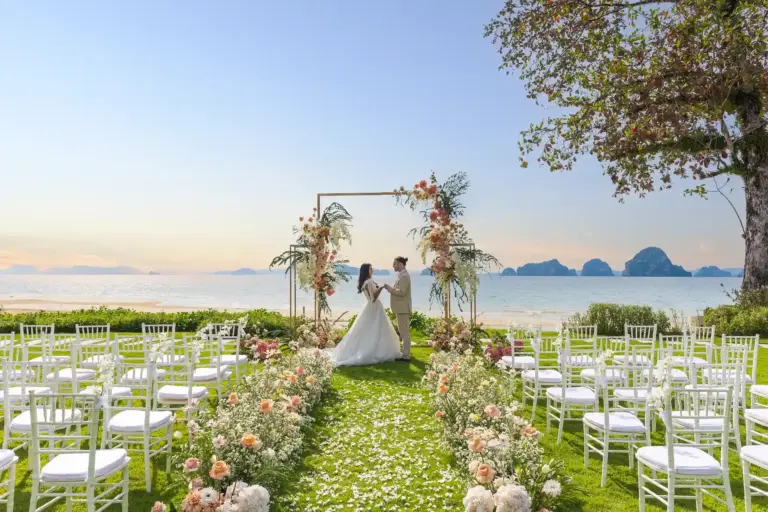 Ein Paar kurz vor der Trauung am thailändischen Strand, im Hintergrund ikonische Felsformationen über dem Meer, im Vordergrund weiße Stuhlreihen und zarter Blütenschmuck. Katharina Fenners ist Ihre Spezialistin für Trauungen und Flitterwochen in Thailand!