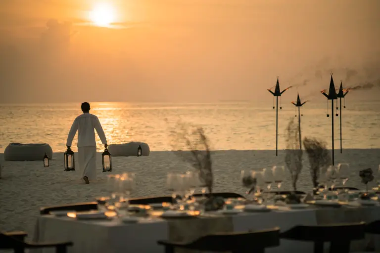 Ein Angestellter eines Privatinsel-Resorts trägt Lampen an den einsamen Strand, im Vordergrund eine festlich gedeckte Tafel - eine Vielzahl maßgeschneiderter Erlebnisse warten im Privatinsel-Urlaub auf Sie, buchbar bei Fenners Reisen