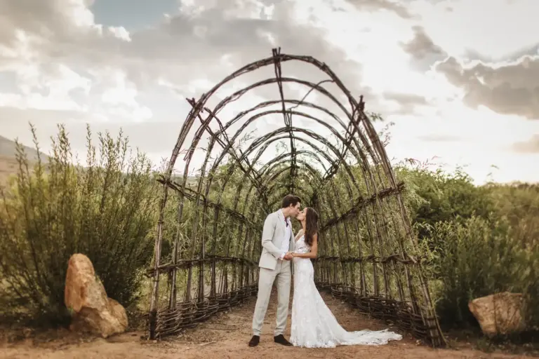 Ein Brautpaar unter einem rustikalen Holzbogen in der blühenden Natur Südafrikas. Ob Victoria Falls oder Safaris mit abenteuerlichen Tierbegegnungen, eine Trauung oder Hochzeitsreise in Afrika bleibt lange in Erinnerung! Katharina Fenners berät Sie individuell.