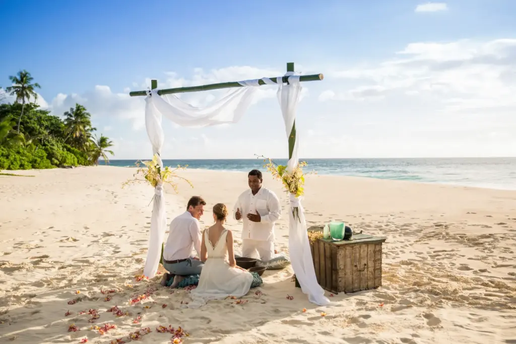 Ein Brautpaar kniet während der Trauung im Sand eines einsamen Seychellen-Strands - die Trauminseln im Indischen Ozean bieten sich für unvergessliche Hochzeiten oder Hochzeitsreisen an! Reise-Expertin Katharina Fenners ist bestens vernetzt vor Ort und organisiert Ihre maßgeschneiderte Hochzeitsreise oder Trauung!