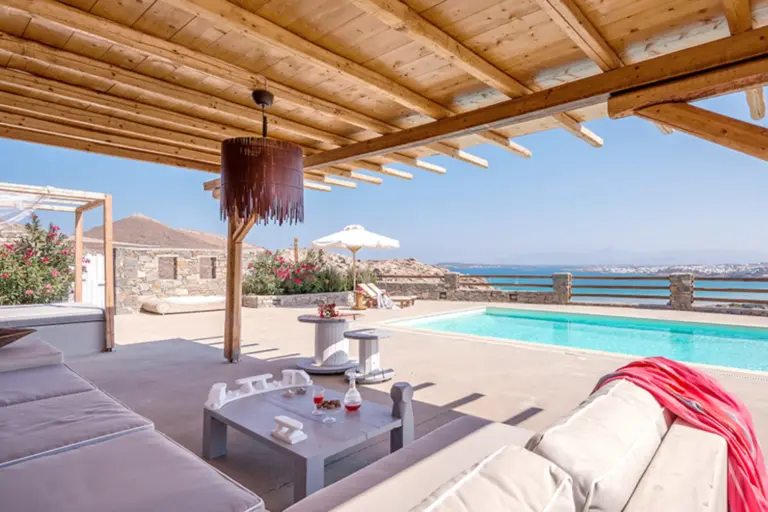 Blick von einem gemütlichen Outdoor-Sofa über den eigenen Pool und das angrenzende Meer - diese Villa auf Griechenland liegt nur wenige Minuten von der mykenischen Akropolis entfernt und verzaubert durch sanfte Erdtöne und mediterrane Eleganz - buchbar bei Fenners Reisen