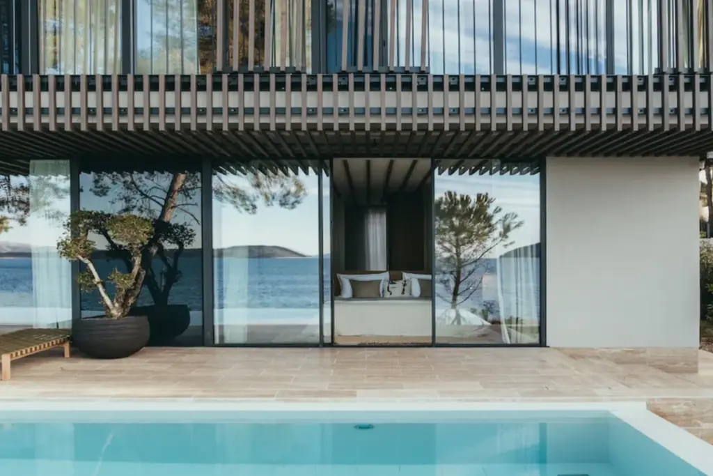 Durch deckenhohe Schiebetüren erhascht man hier einen Blick ins gediegene Schlafzimmer einer Luxus-Ferienvilla, im Vordergrund der Pool. Jetzt bei Fenners Reisen Ihre Traumvilla mieten, für intime Ferientage auf höchstem Niveau!