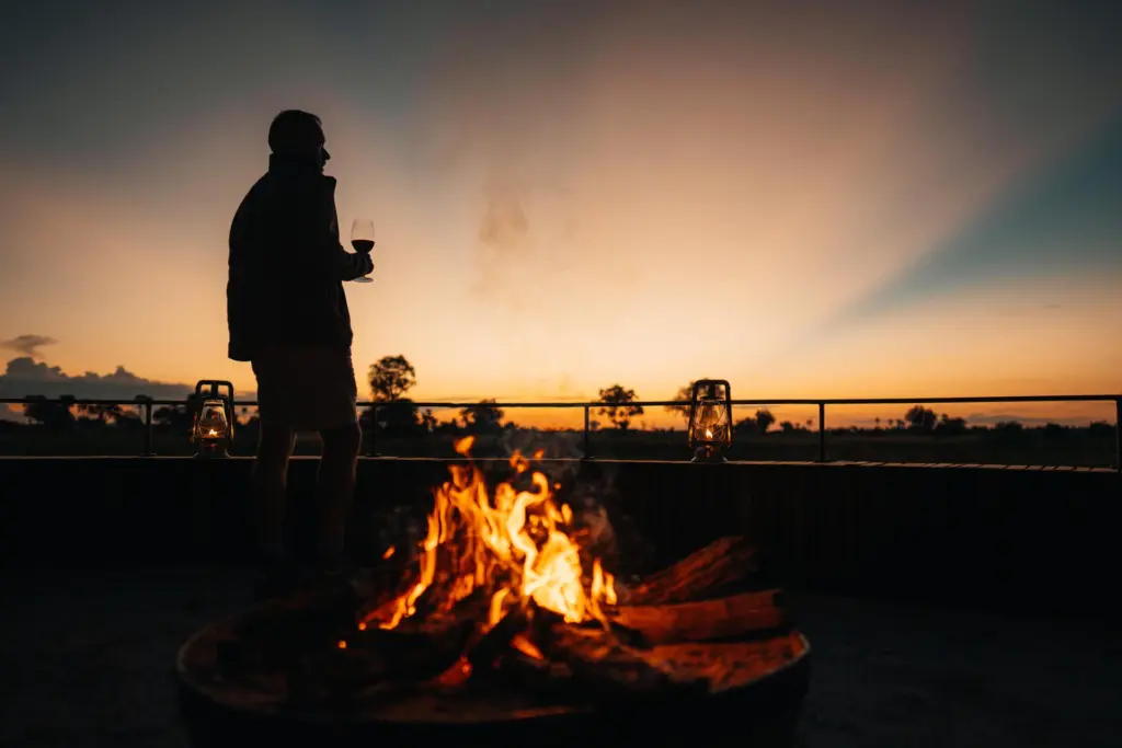 Ein Mann genießt ein Glas Wein vor einem flackernden Feuer, im Hintergrund dehnt sich die weite Savanne in der Dämmerung. Botswana-Expertin Katharina Fenners kennt die exklusivsten Camps und Lodges für Ihre einzigartige Botswana Safari!