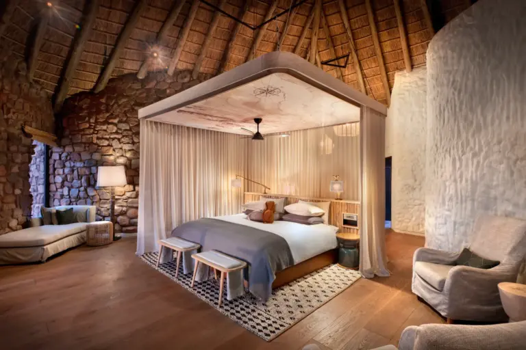 Ein Himmelbett in einem hohen, gemäuerten Raum mit rustikalem Dekor und modernen Akzenten - die eleganten, strohgedeckten Suiten von Tswalu Motse bieten viel Platz und atemberaubende Aussichten auf die Kalahari. Katharina Fenners organisiert Ihren maßgeschneiderten Aufenthalt!