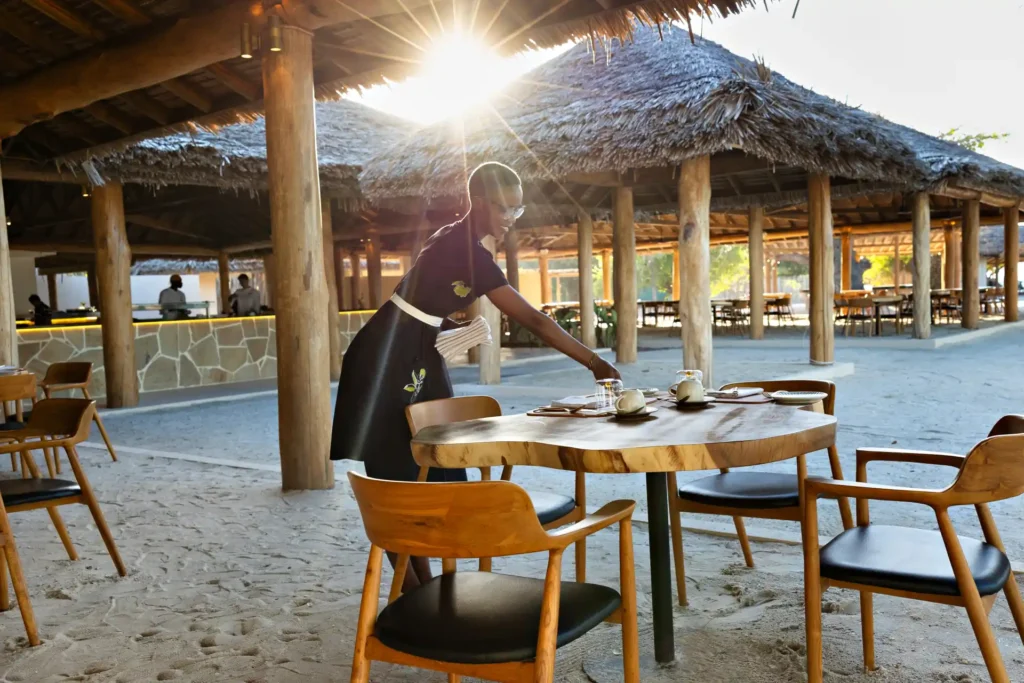 Edle Holztische und Stühle im Sand werden von einer freundlichen Kellnerin gedeckt - auf Bawe Island Zanzibar stimmt jedes Detail. Katharina Fenners ermöglicht Ihren individuellen Aufenthalt!
