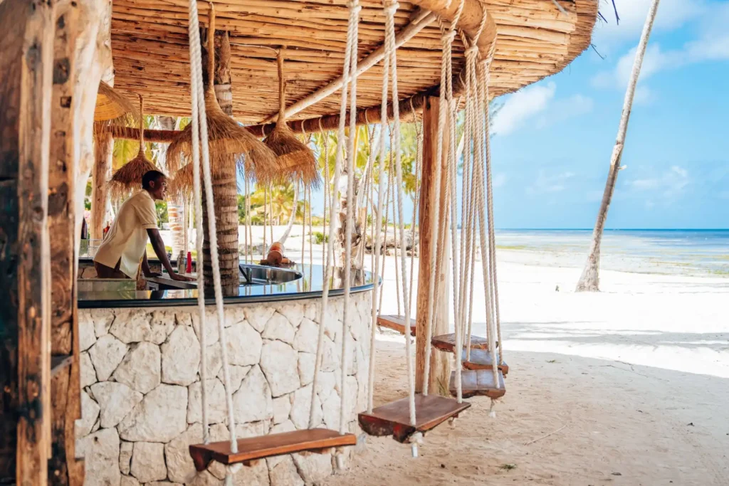 Gut gelaunter Kellner in einer Strandbar mit Schaukeln statt Stühlen und paradiesischem Strand im Hintergrund. Das ist Sansibar - buchbar nach Maß bei Fenners Reisen!