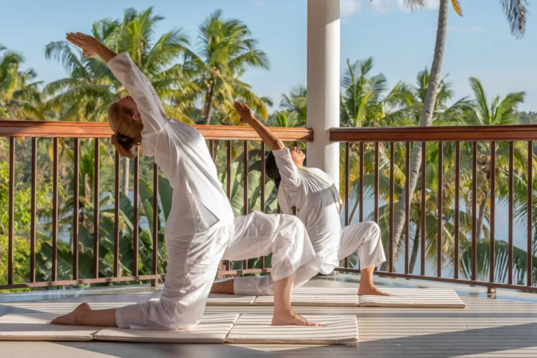 Zwei Frauen praktizieren Yoga auf einer Terrasse mit Blick auf Kokospalmen und einen See. Neben Ernährung spielen auch Yoga und Meditation eine große Rolle im ayurvedischen Heilungsprozess. Buchen Sie eine Gesundheitsreise nach Indien bei Fenners Reisen!