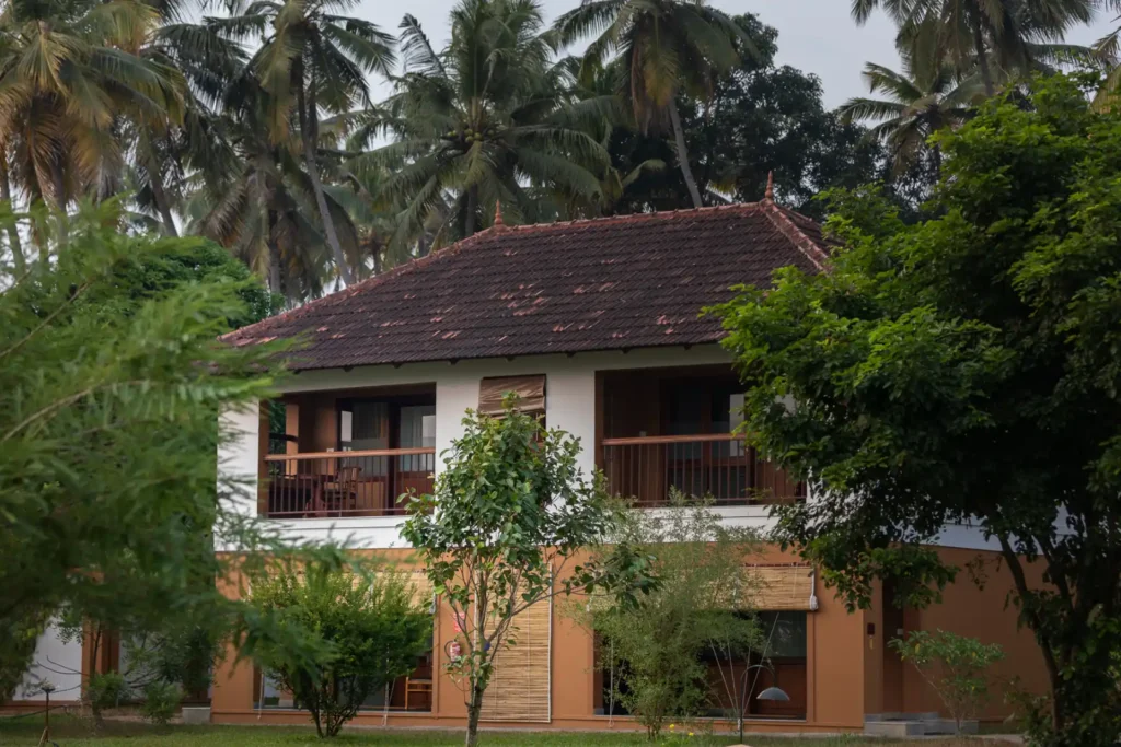 Zweistöckiges Gebäude zwischen Palmen. Die Anlage der Kalari Rasayana Ayurvedaklinik erstreckt sich auf einem Areal von acht Hektar und liegt ruhig an einem See. Buchen Sie Ihre maßgeschneiderte Gesundheitsreise nach Indien bei Fenners Reisen!