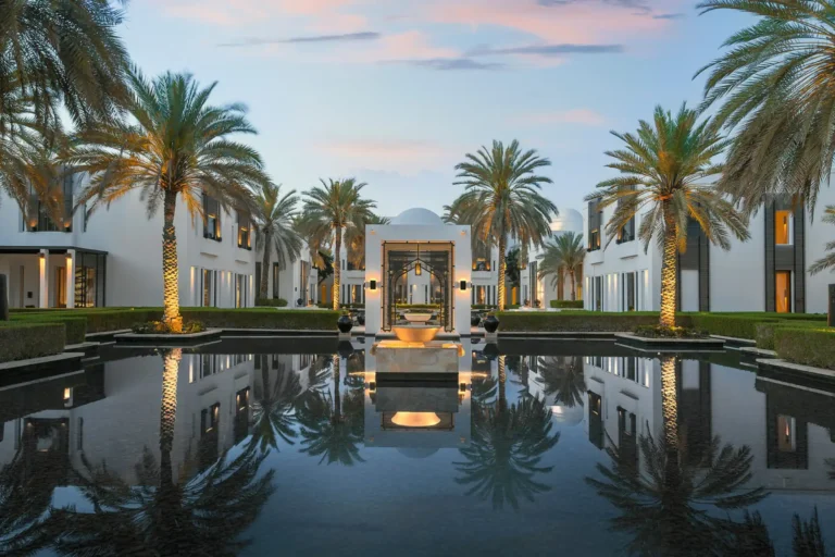 Die streng symmetrischen Gebäude und Palmen des The Chedi Muscat im Oman spiegeln sich im Wasserbecken - wer nach der Anreise in den Oman efrischende Entspannung sucht, ist hier richtig! Buchbar für Ihre individuelle Oman Reise bei Fenners Reisen.