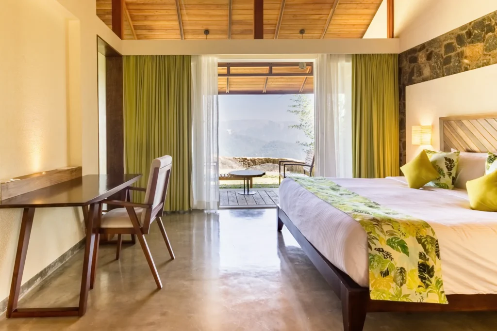 Schreibtisch und Bett in einem Zimmer mit hohen Decken, hinter der bodentiefen Glasfront ein Balkon mit spektakulärem Talblick - dieses Zimmer können Sie bei Fenners Reisen für Ihren Gesundheitsurlaub in Indien buchen!