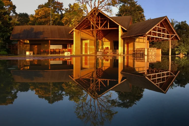 Blick auf ein ikonisch geschnittenes Gebäude des Prakriti Shakti vor einem Pool im goldenen Sonnenlicht - die Architektur dieses Gesundheitsresorts in Indien besticht durch ihre dezente Natürlichkeit. Buchbar ist der Aufenthalt ganz individuell bei Fenners Reisen!