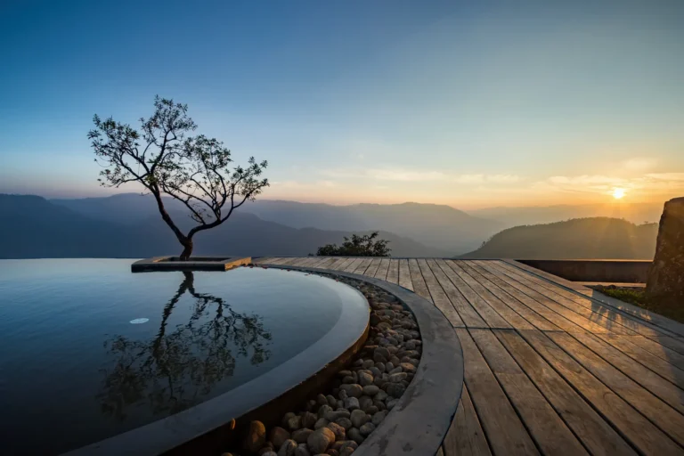 Der geschwungene Infinity Pool des Praktiki Shakti mit Blick auf die spektakuläre Hügellandschaft Keralas im Sonnenuntergang. Buchbar ist der Aufenthalt in der Gesundheitsklinik in Indien bei Fenners Reisen.