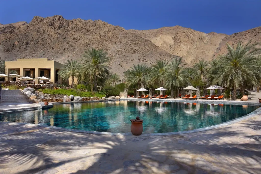 Poollandschaft, Palmen und niedrige Lehmgebäude, im Hintergrund hohe Berge -eine Kulisse wie aus Tausendundeiner Nacht bietet sich Gästen des Six Senses Zighy Bay, buchbar bei Fenners Reisen