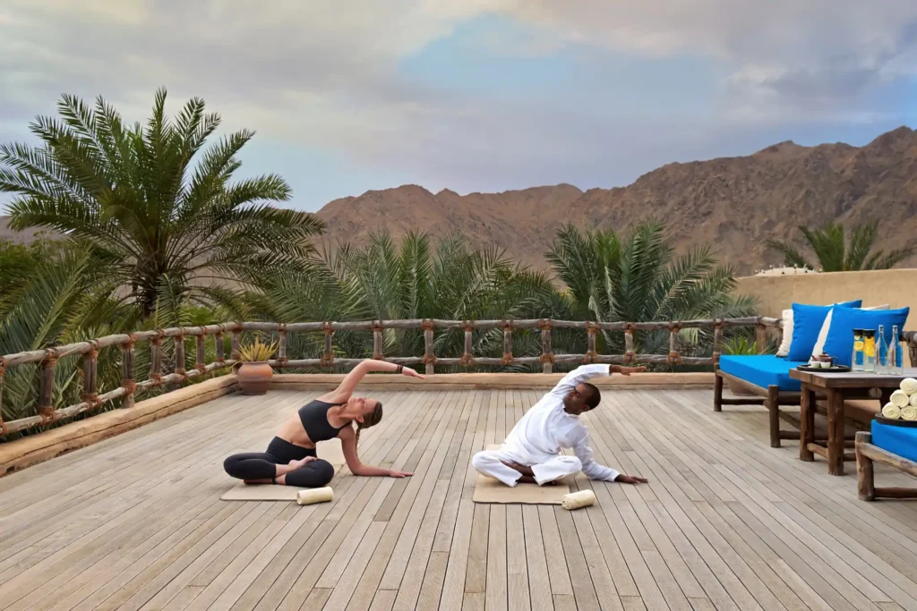 Yogastunde auf einer Terrasse zwischen den Palmkronen, im Hintergrund das Omanische Gebirge - das Six Senses Zighy Bay bietet viel Abwechslung! Buchen Sie eine unvergessliche Oman Reise bei Fenners Reisen.