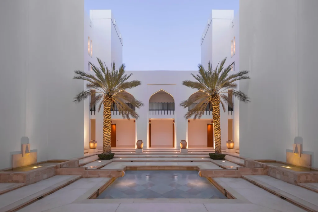 Symmetrische Gebäude aus hellem Stein vor einem Wasserbecken - das archtektonisch preisgekrönte The Chedi Muscat bietet Ruhe und Erholung nach der Anreise, und zugleich die Möglichkeit das Umland zu erkunden! Katharina Fenners gestaltet Ihre individuelle Reise in den Oman.