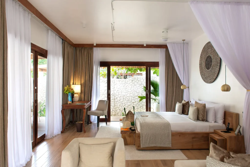 Luftige Vorhänge, bodentiefe Fenster und natürliche Materialien dominieren in diesem Schlafzimmer einer Garten-Villa mit Whirlpool, buchbar für einen Sansibar Urlaub an einem Bilderbuchstrand bei der Expertin Katharina Fenners.